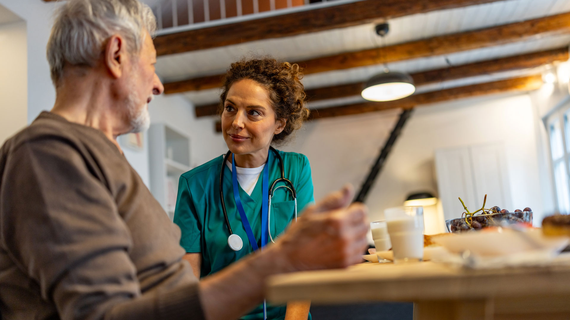 A caring nurse in green scrubs chats with an elderly man at a home dining table, fostering trust and comfort during a meal. QQFS. Case study. Malnutrition.